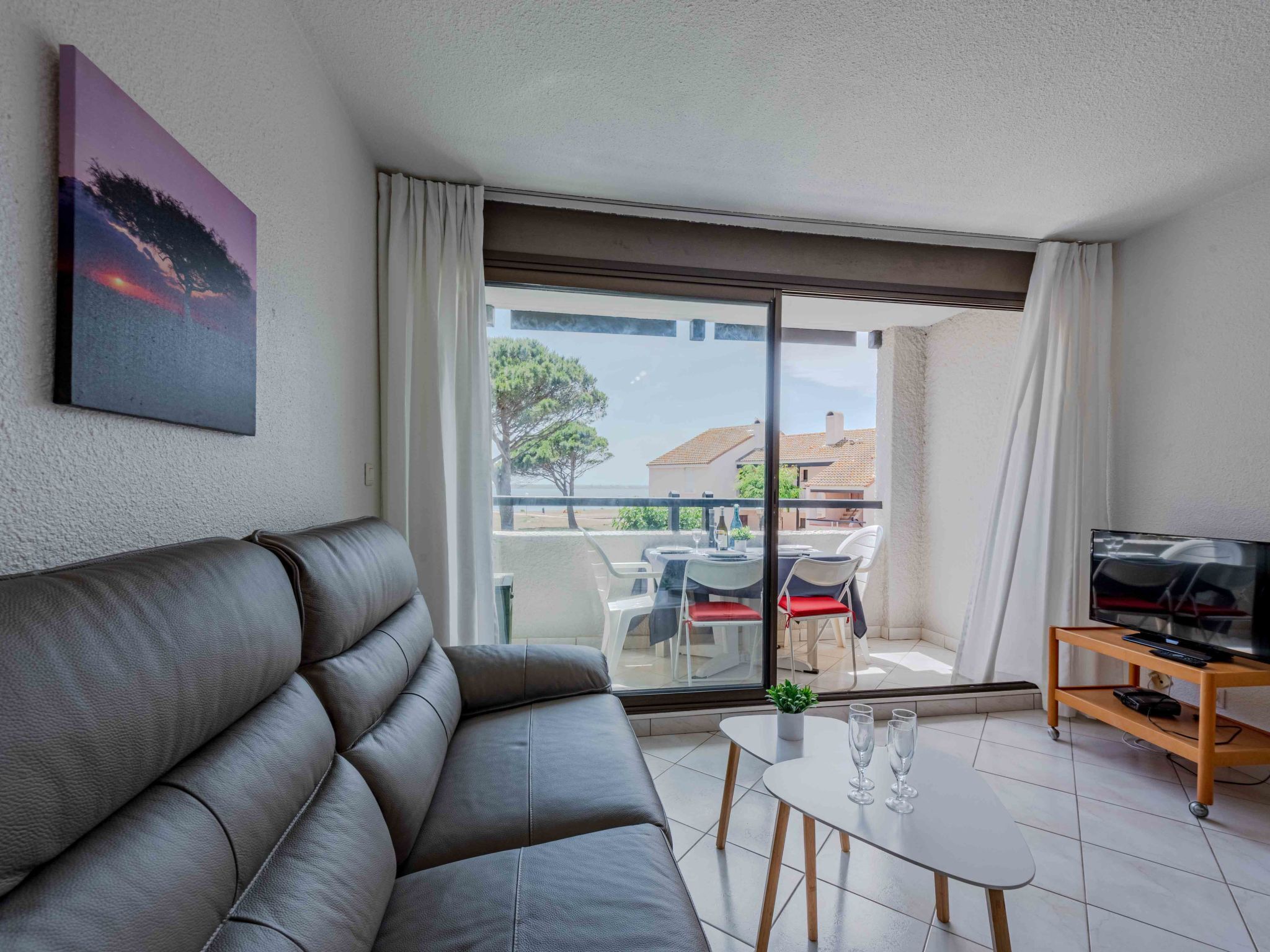 Una sala de estar moderna con un sofá de cuero, una mesita con vasos, un televisor sobre un mueble de madera y un balcón con vistas al mar y edificios.