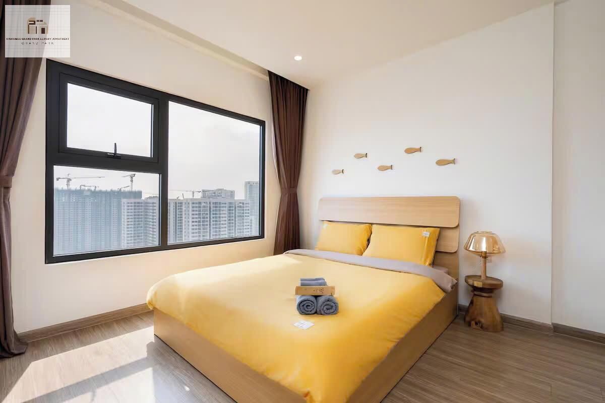 A hotel room with a large window, a bed with yellow bedding, a wooden headboard, and a small bedside table with a lamp.