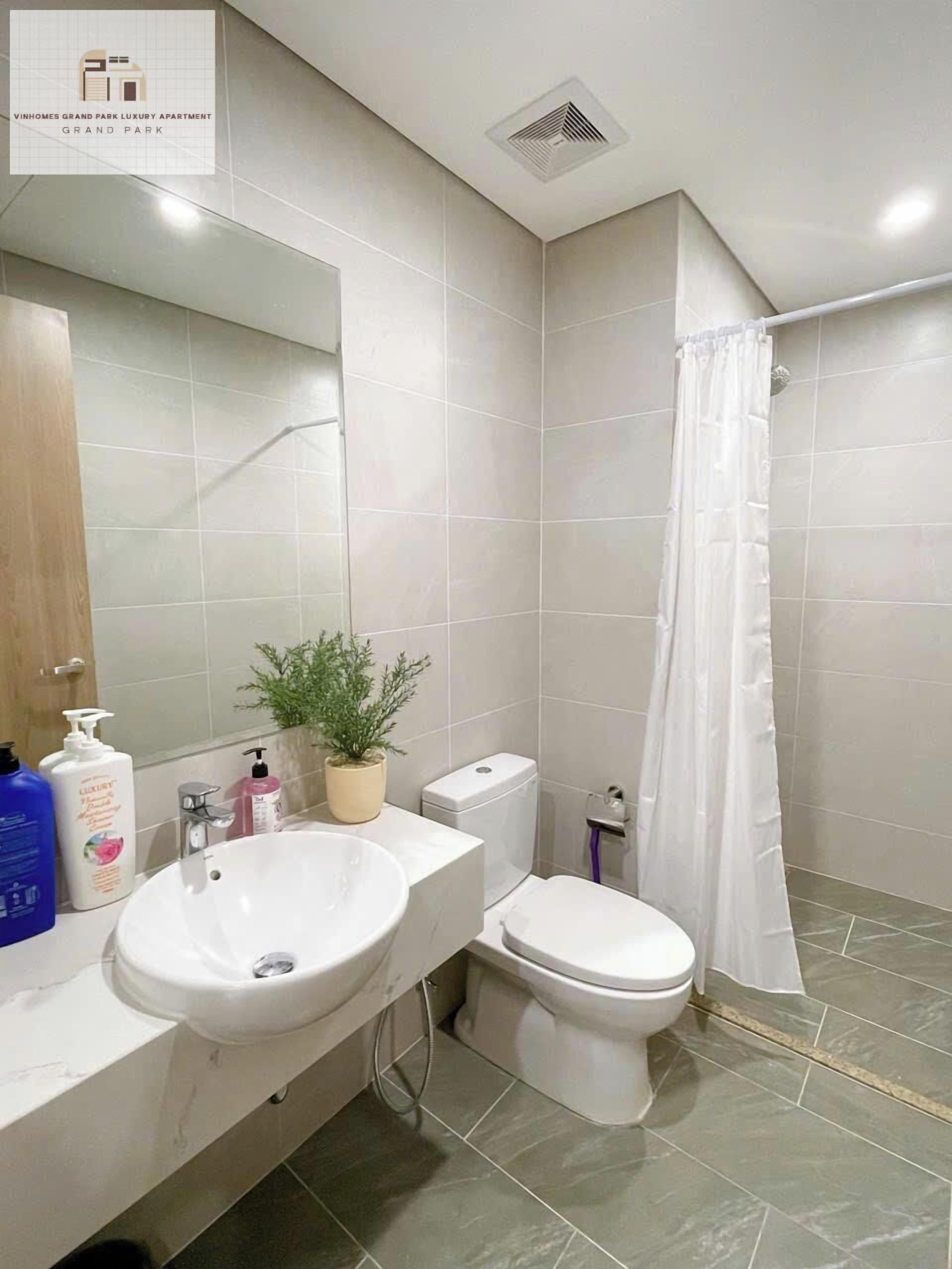 Un baño con lavamanos, inodoro y cortina de ducha blancos. Hay un espejo sobre el lavamanos y una planta en maceta en la encimera.