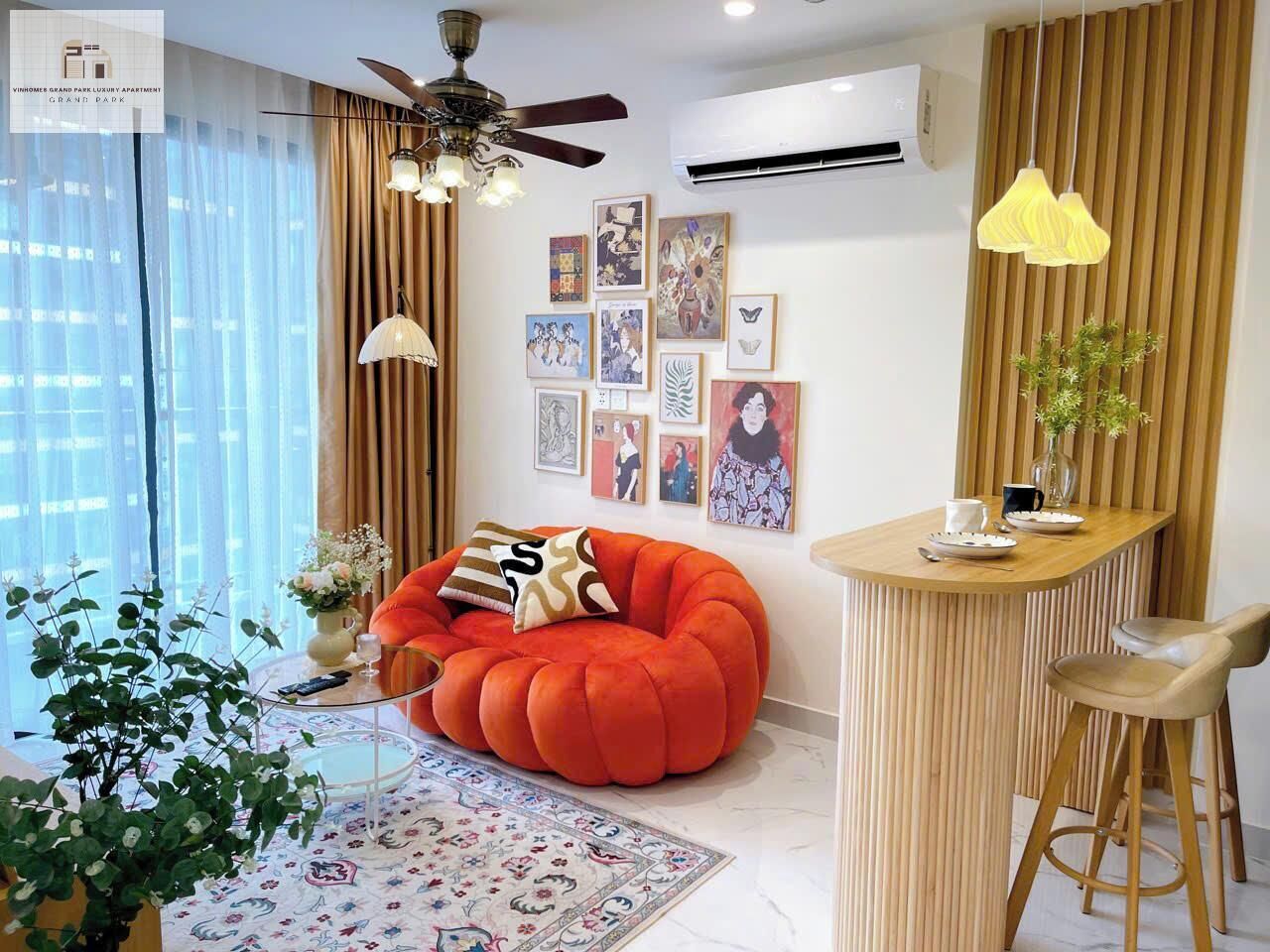 A modern living room with a round orange bean bag, a wooden dining table, and a small bar stool. There are framed artworks on the wall and a ceiling fan above.