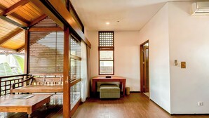 A spacious room with a wooden floor, a wooden bench, a table, and a sliding door leading to an outdoor area. - Villa Uma Pertiwi by Nakula (Seminyak)