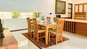 A dining area with a wooden table and chairs, a vase with flowers, and a kitchen with wooden cabinets and a stone backsplash. - Villa Uma Pertiwi by Nakula (Seminyak)