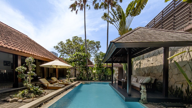 A swimming pool with lounge chairs and umbrellas, surrounded by tropical plants and a stone wall. - Villa Uma Pertiwi by Nakula (Seminyak)