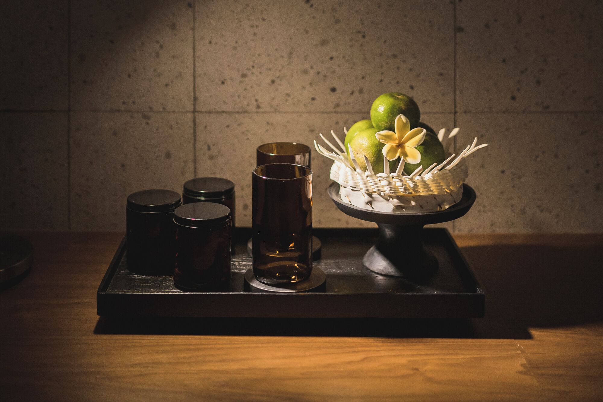 A tray with a decorative arrangement of green apples and a white flower, accompanied by three dark glasses and two small containers.