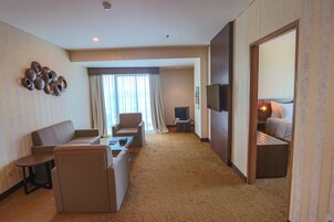 Quarto de hotel com sofá, poltrona, mesa pequena, televisão e cama com mesa de cabeceira.