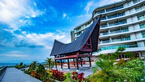 A modern hotel with a distinctive curved roof structure, a covered outdoor area with seating, and lush greenery. - Labersa Hotel And Convention Center Toba Balige (Balige)