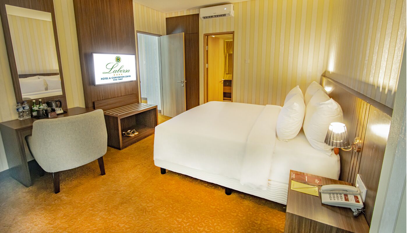 A hotel room with a bed, a desk, a chair, a TV, and a minibar.