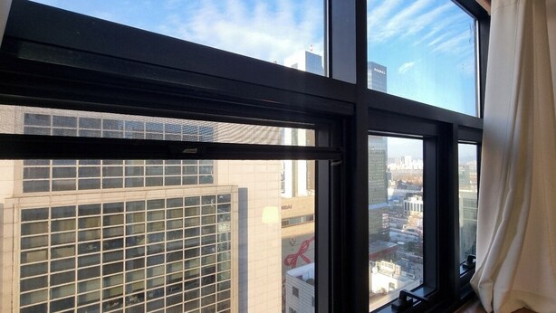 A view through a window with a grid pattern, a curtain, and a glimpse of a cityscape with buildings.