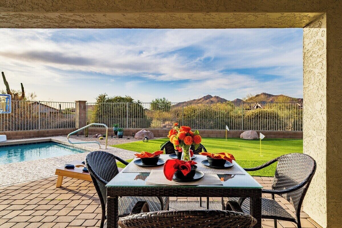Une terrasse avec une piscine, une table à manger et des chaises, donnant sur un jardin avec un drapeau et des montagnes à l’horizon.