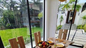 Een ontbijttafel met eten, zoals toast, eieren en fruit, voor een groot raam met uitzicht op een tuin met palmbomen.