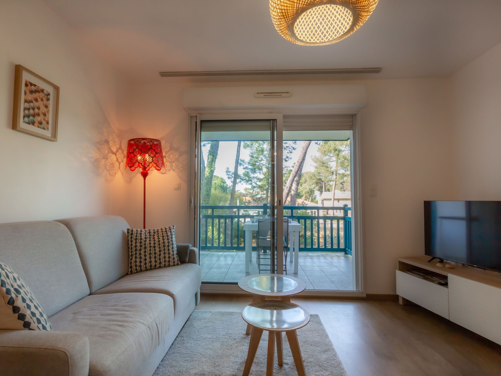 A living room with a sofa, a coffee table, a TV, and a balcony with a view of trees and a fence.