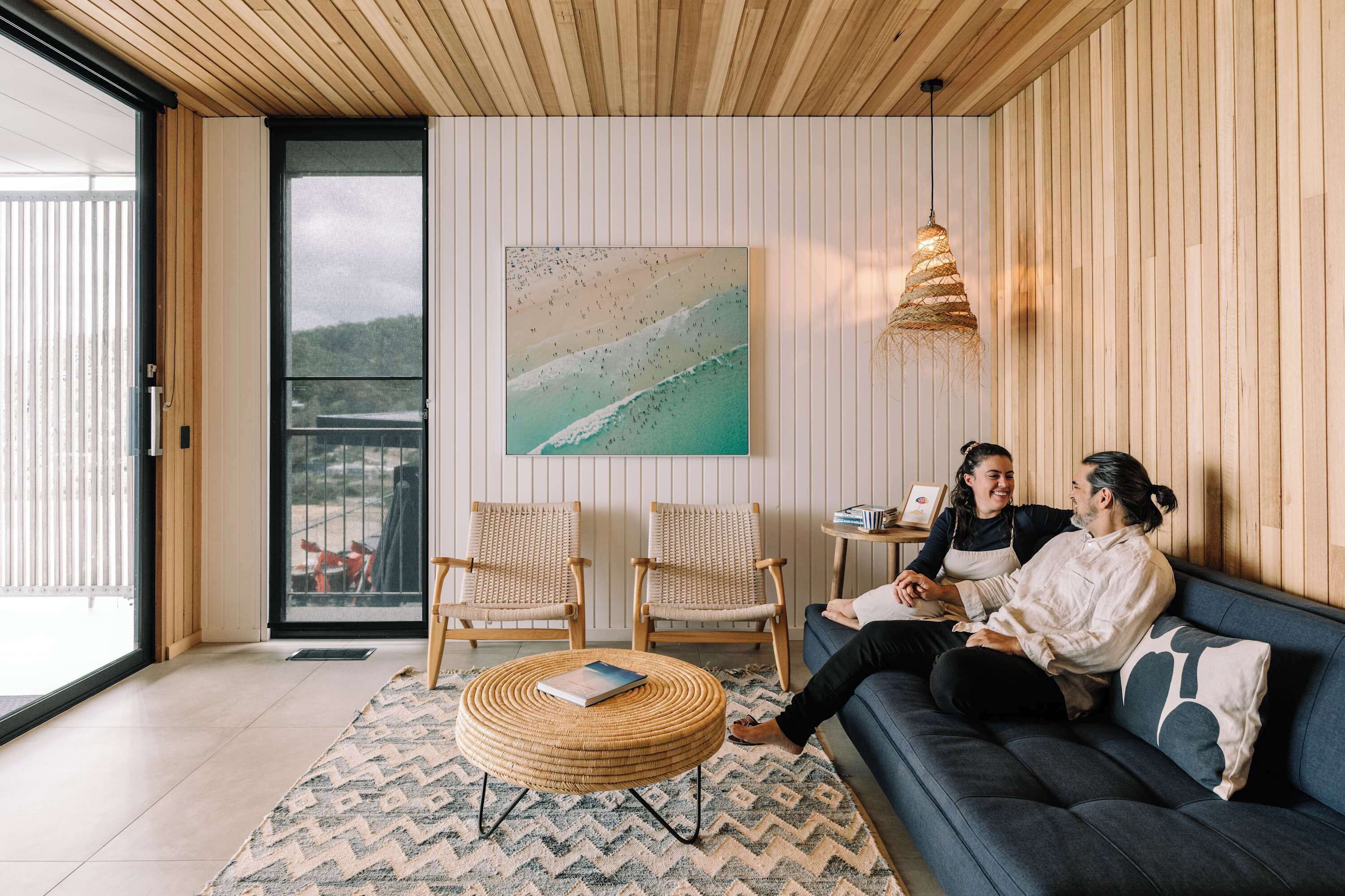 A modern living room with a sofa, two wicker chairs, a round coffee table, a painting, and a hanging light fixture.