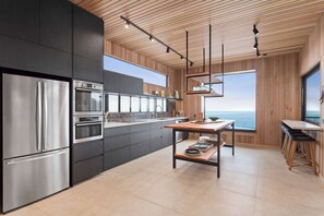 A modern kitchen with stainless steel appliances, a wooden ceiling, and a view of the sea.