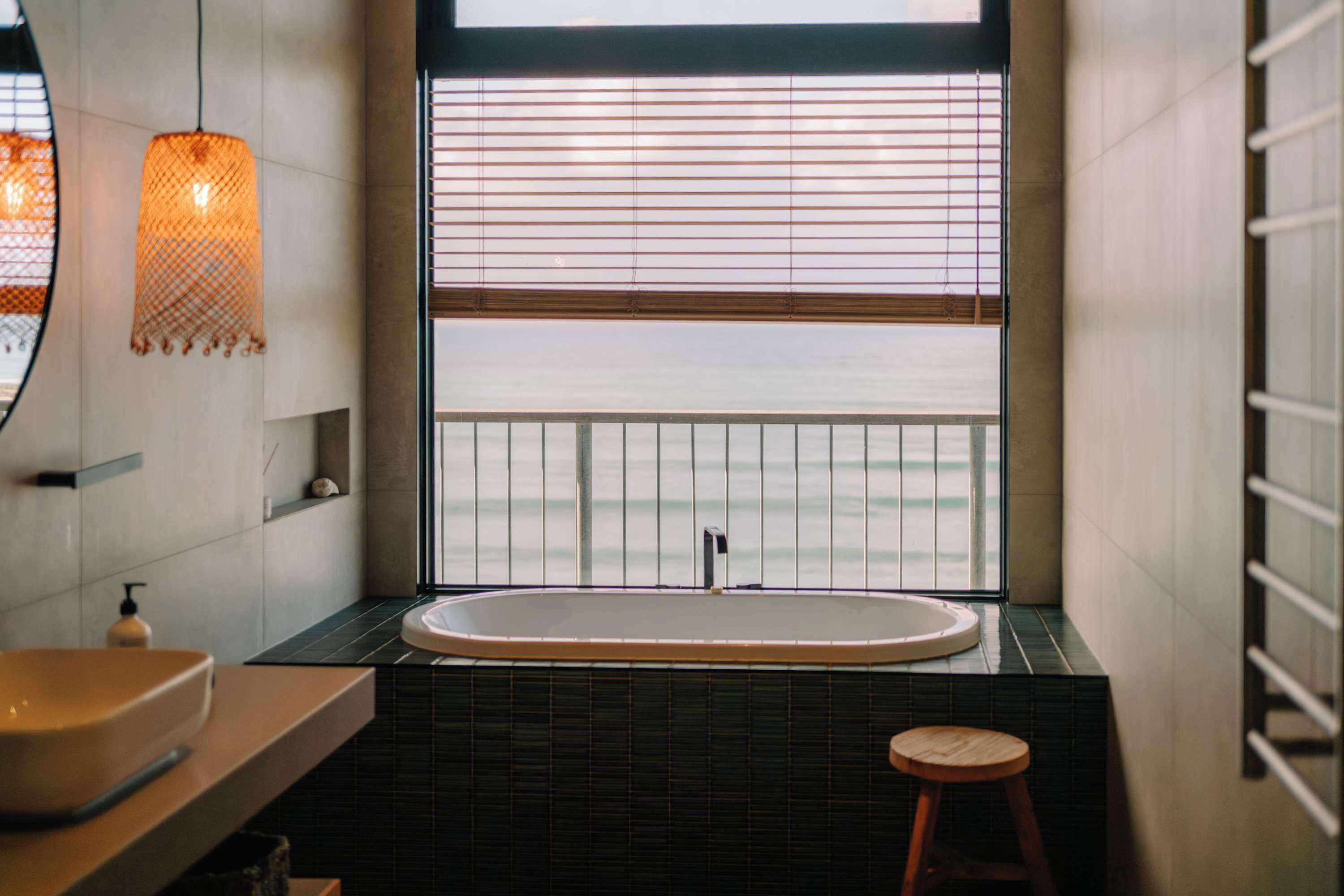 A modern bathroom with a freestanding bathtub, a wooden stool, and a large window with blinds.