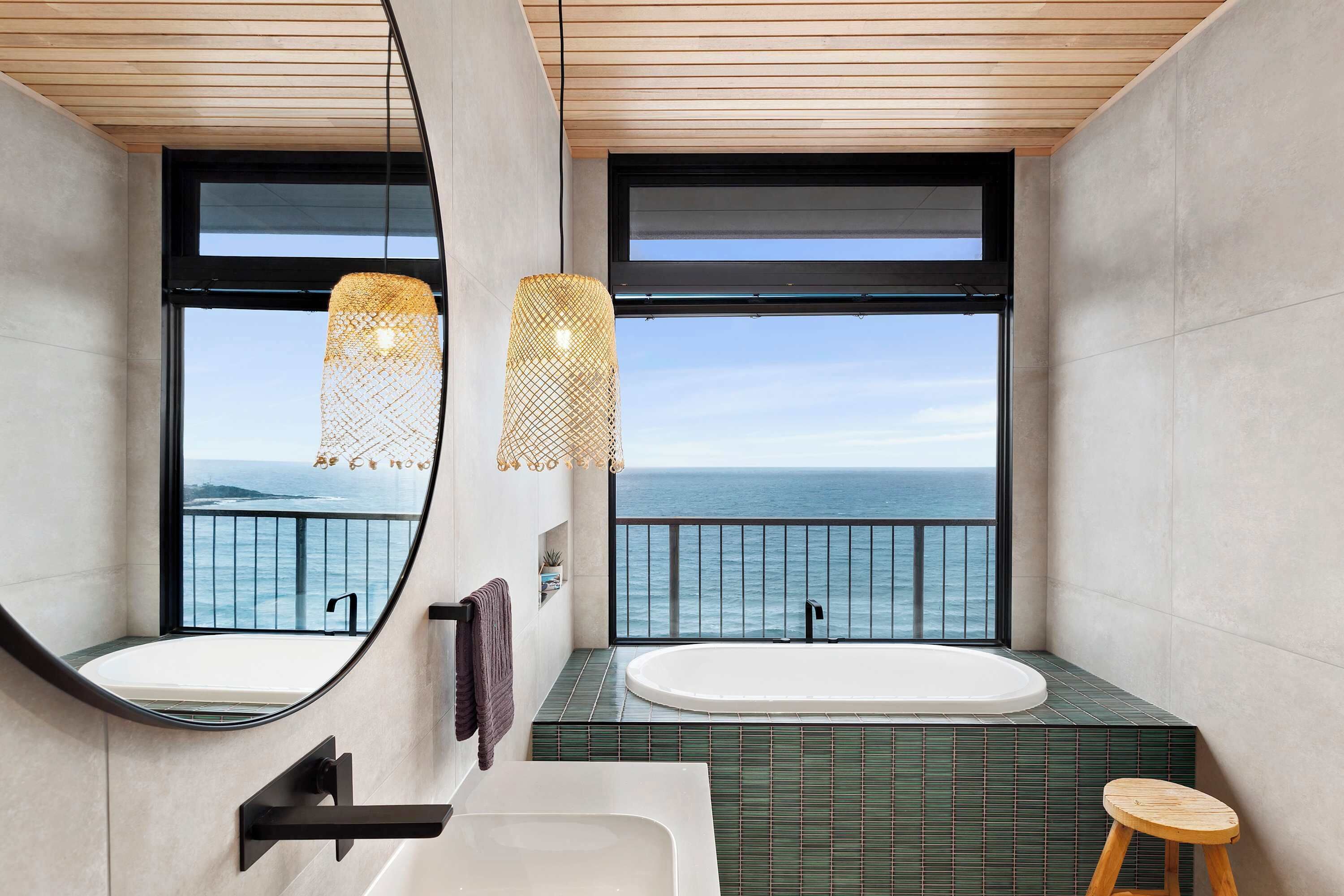 A modern bathroom with a large bathtub, a round mirror, and a view of the ocean.