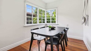 A dining area with a dark wooden table and chairs, a window with a view of greenery, and a white wall with a power outlet.