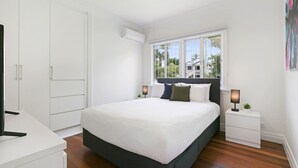 A bedroom with a large bed, a wardrobe, a bedside table with a lamp, and a window with a view of buildings.
