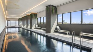 A modern indoor swimming pool with clear glass walls, lounge chairs, and a view of the landscape. - High-Rise 3-Bed Apartment with Premium Amenities (Box Hill)