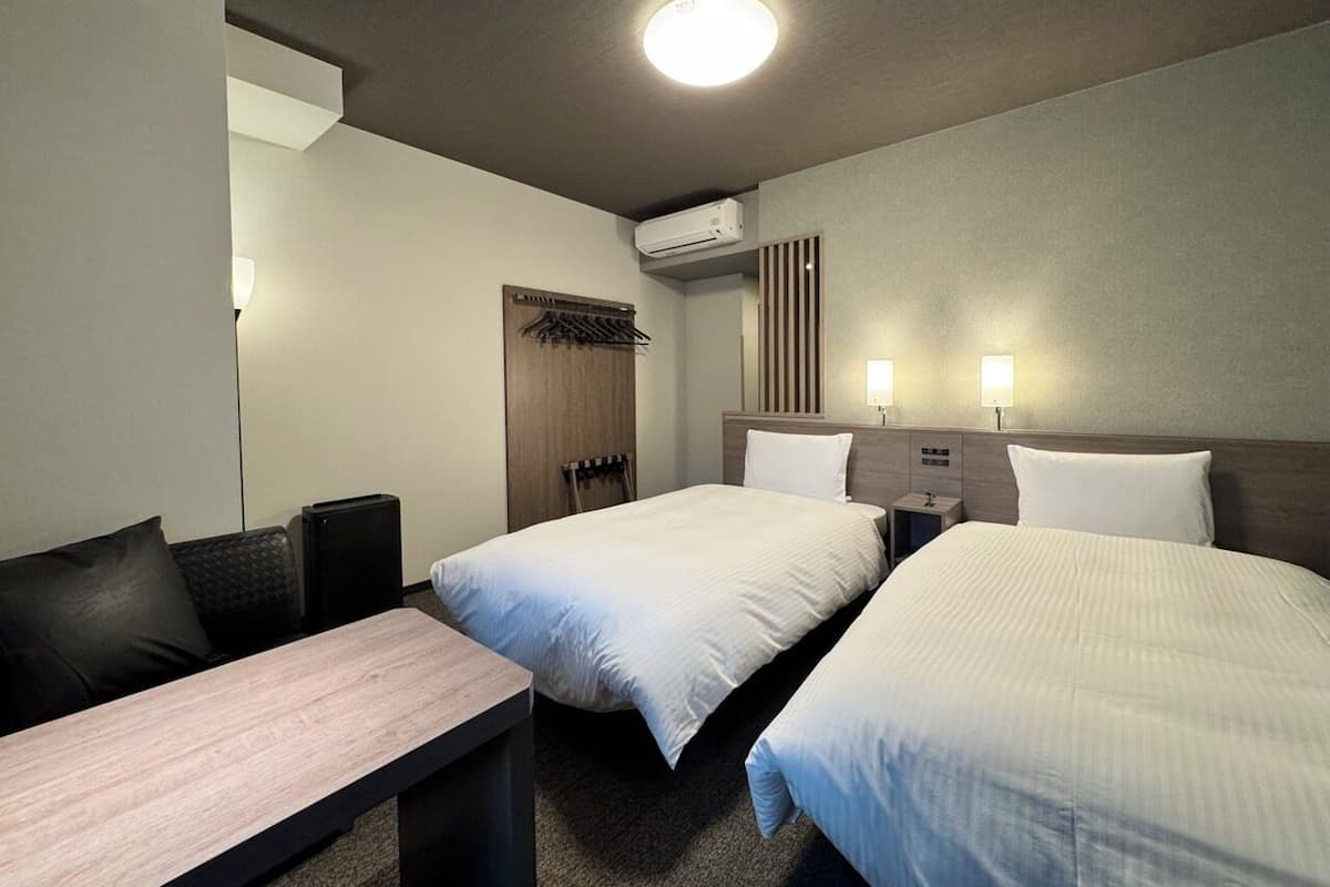 Habitación de hotel con dos camas, un escritorio, una silla, una mesita pequeña, una mesita de noche y un equipo de aire acondicionado.