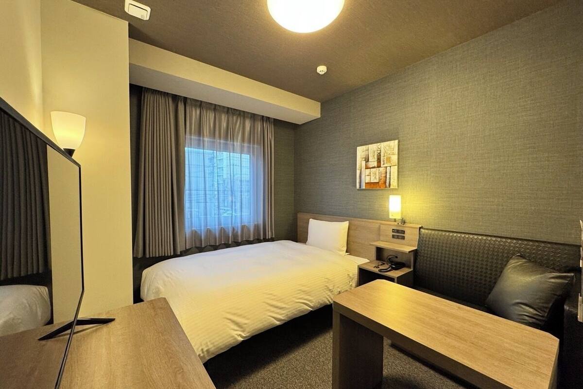 Habitación de hotel con cama, escritorio, silla, mesita de noche y lámpara de pared.