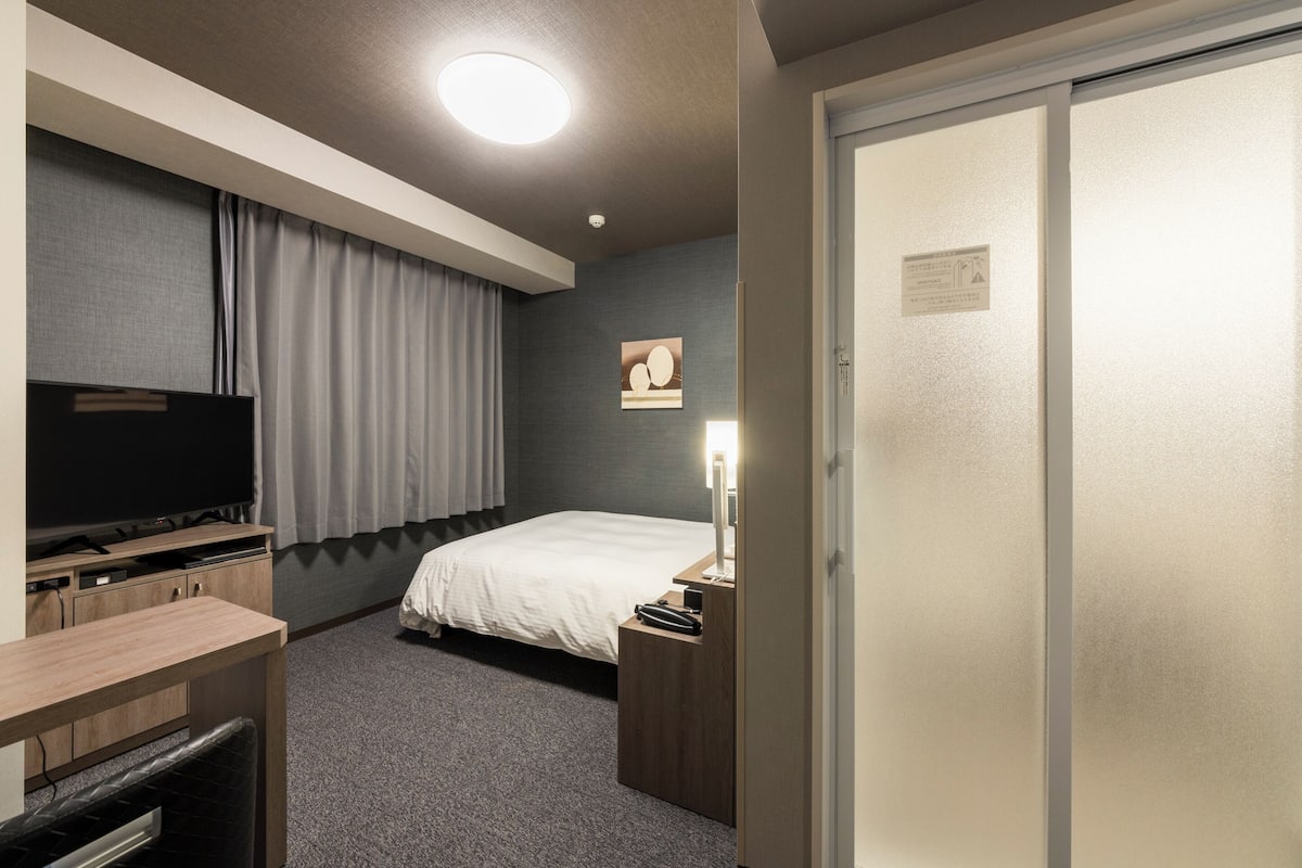 Una habitación de hotel con cama, escritorio con televisor, mesita de noche con lámpara y armario.