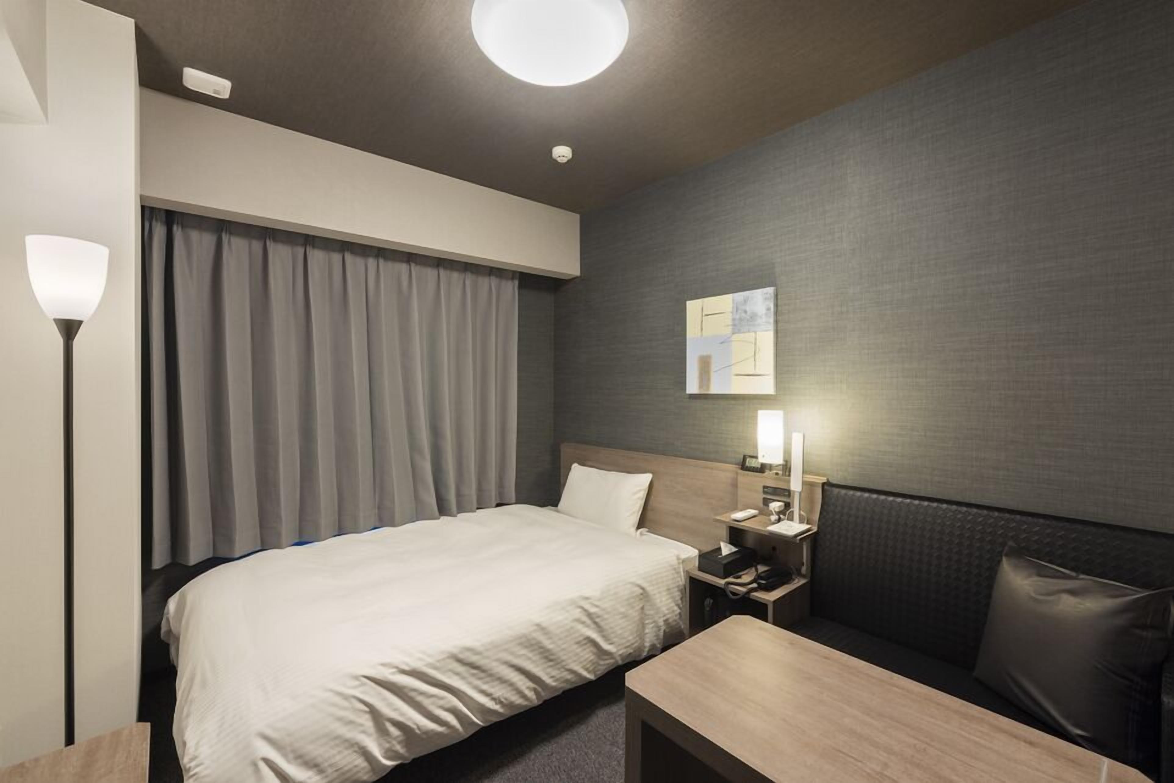 A modern hotel room with a bed, bedside table, lamp, and a small seating area.