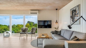 A modern living room with a grey sofa, a round coffee table, and two furry chairs. A TV is mounted on the wall, and there's a large window offering a view of greenery. - "Shaya" - 325m from the sea by Interhome (Allingåbro)