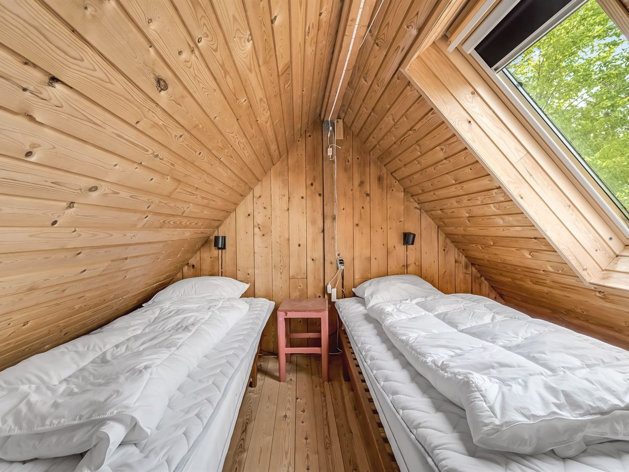 A small, wooden cabin with two single beds, a small table, and a window providing a view of greenery outside.