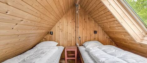 A small, wooden cabin with two single beds, a small table, and a window providing a view of greenery outside.