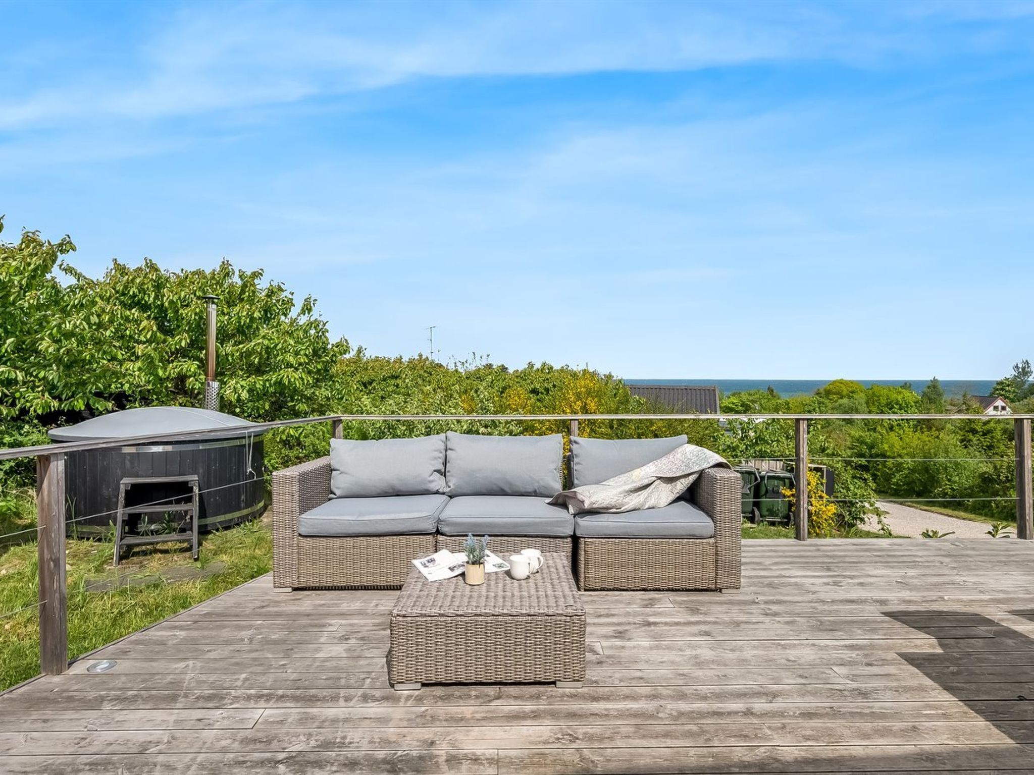 A wooden deck with a wicker sofa and a small table, surrounded by greenery and a clear sky.