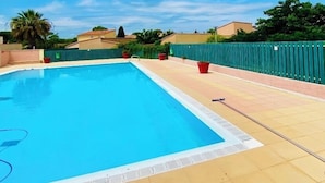 Una piscina rectangular con solárium de baldosas, rodeada por una cerca verde, bajo un cielo azul despejado.