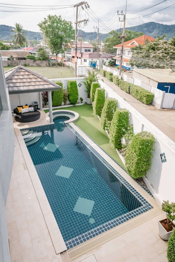 A swimming pool with a tiled deck, a covered seating area, and a view of a residential neighborhood.