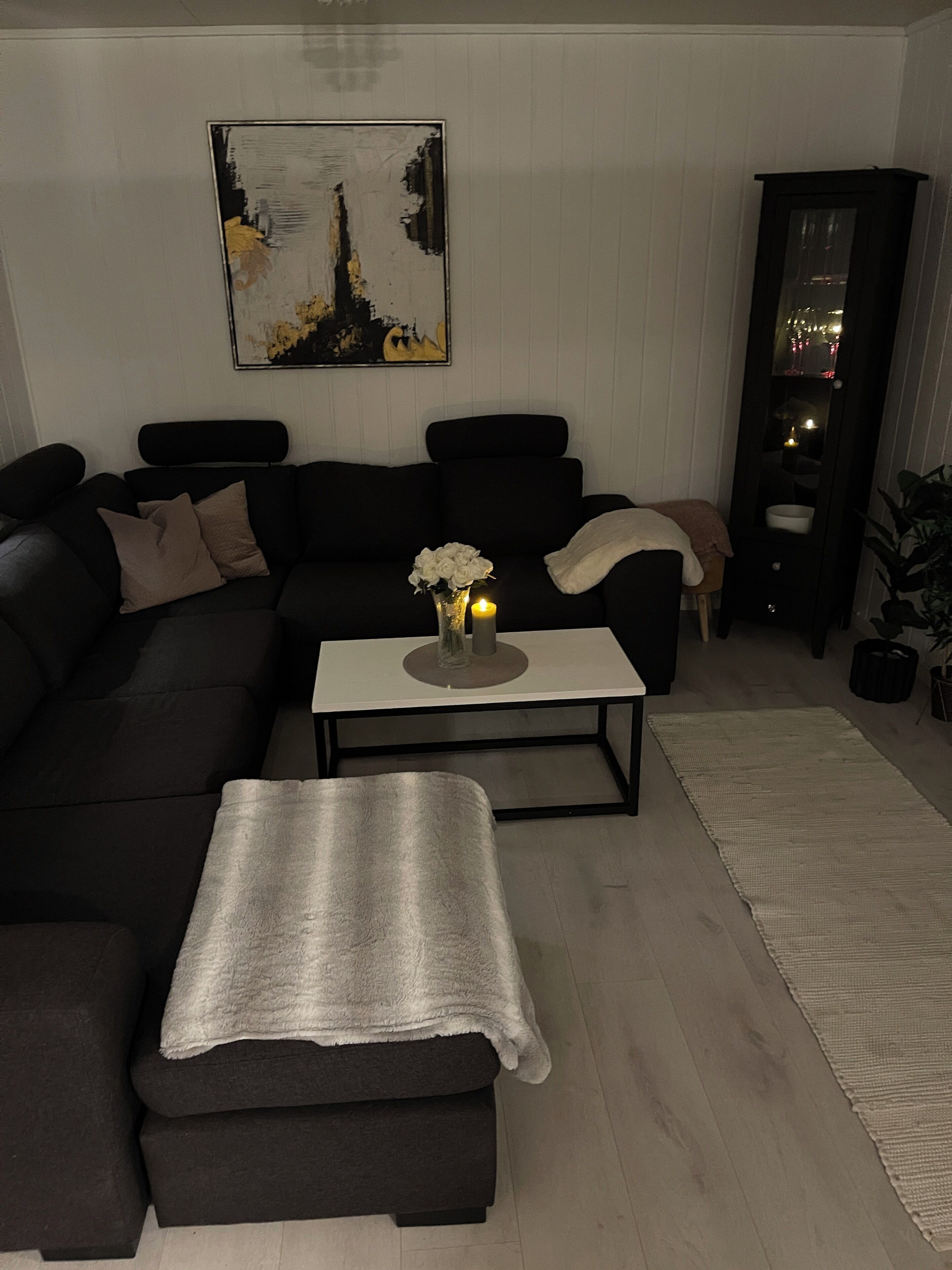 Una sala moderna con un sofá seccional, una mesita de centro con un jarrón de flores blancas y velas, y un cuadro en la pared.