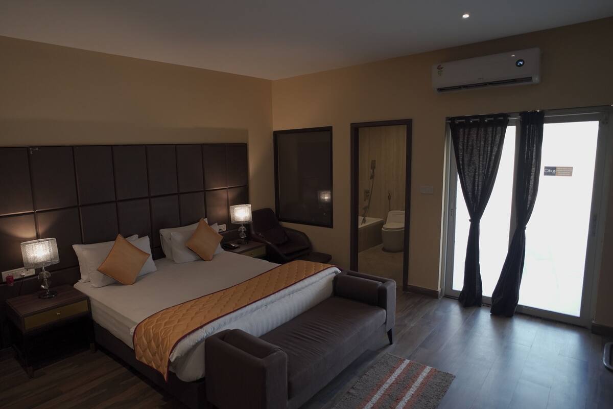 A modern hotel room with a large bed, bedside tables, a desk, a chair, and a view of the outside through a window with curtains.