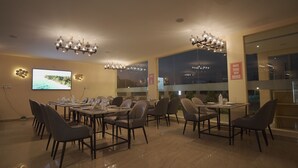 A modern dining area with multiple tables and chairs, a large screen displaying a scenic image, and a view of the city at night. - Citrus Classic Resorts & Spa Mahabalipuram ECR (Cheyyur)