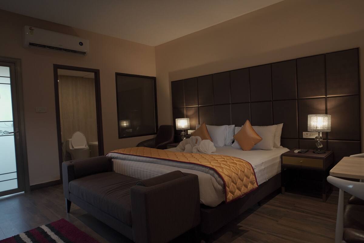 A hotel room with a large bed, a bedside table with a lamp, a chair, a desk, and a window.