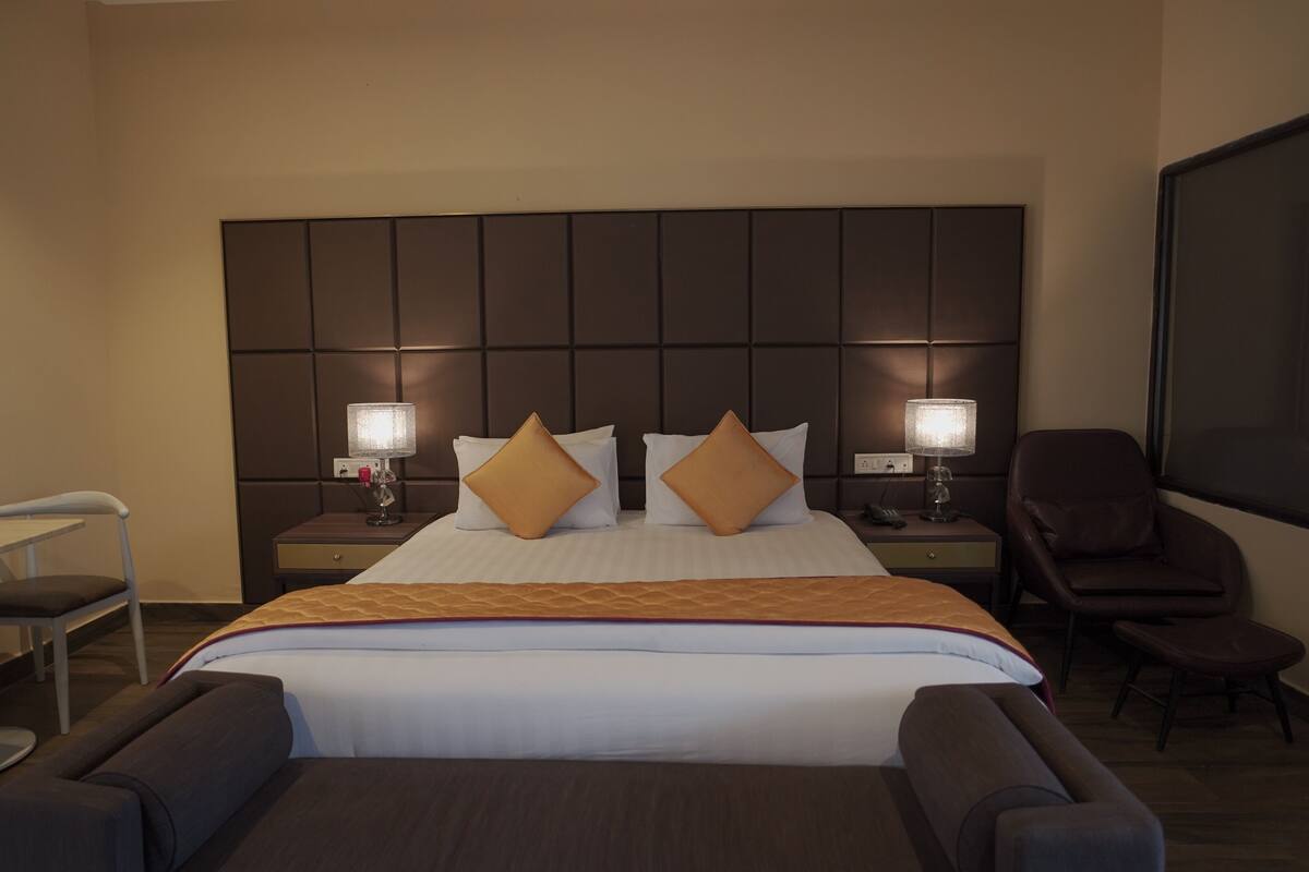A hotel room with a large bed, two bedside tables with lamps, a desk, a chair, and a TV screen.