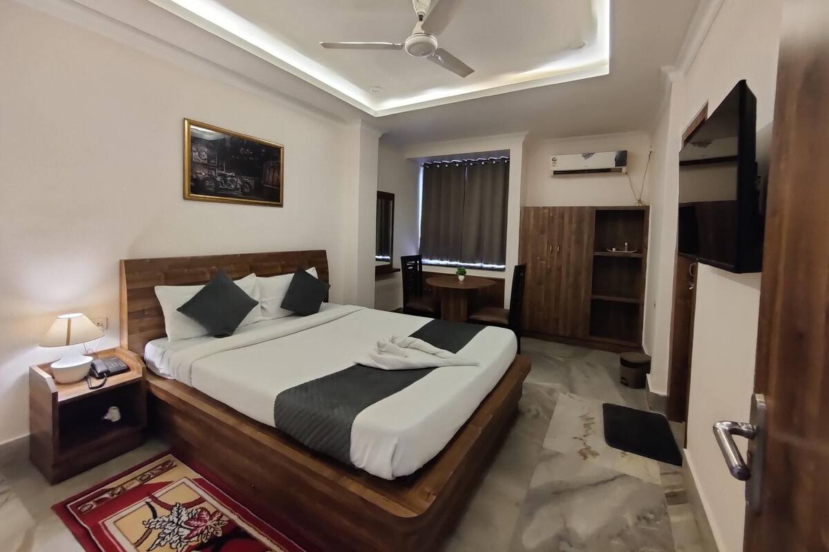 A modern hotel room with a wooden bed, a ceiling fan, a bedside table with a lamp, a small round table, and a large mirror.
