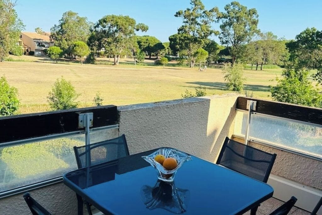 A balcony with a table and chairs, overlooking a grassy area with trees and a building in the background.