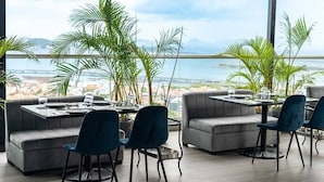A modern dining area with grey sofas and blue chairs, large windows offering a view of the sea and cityscape.