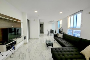 A modern living room with a flat-screen TV, a dark-colored sofa, a glass coffee table, and a dining area with chairs and a table.