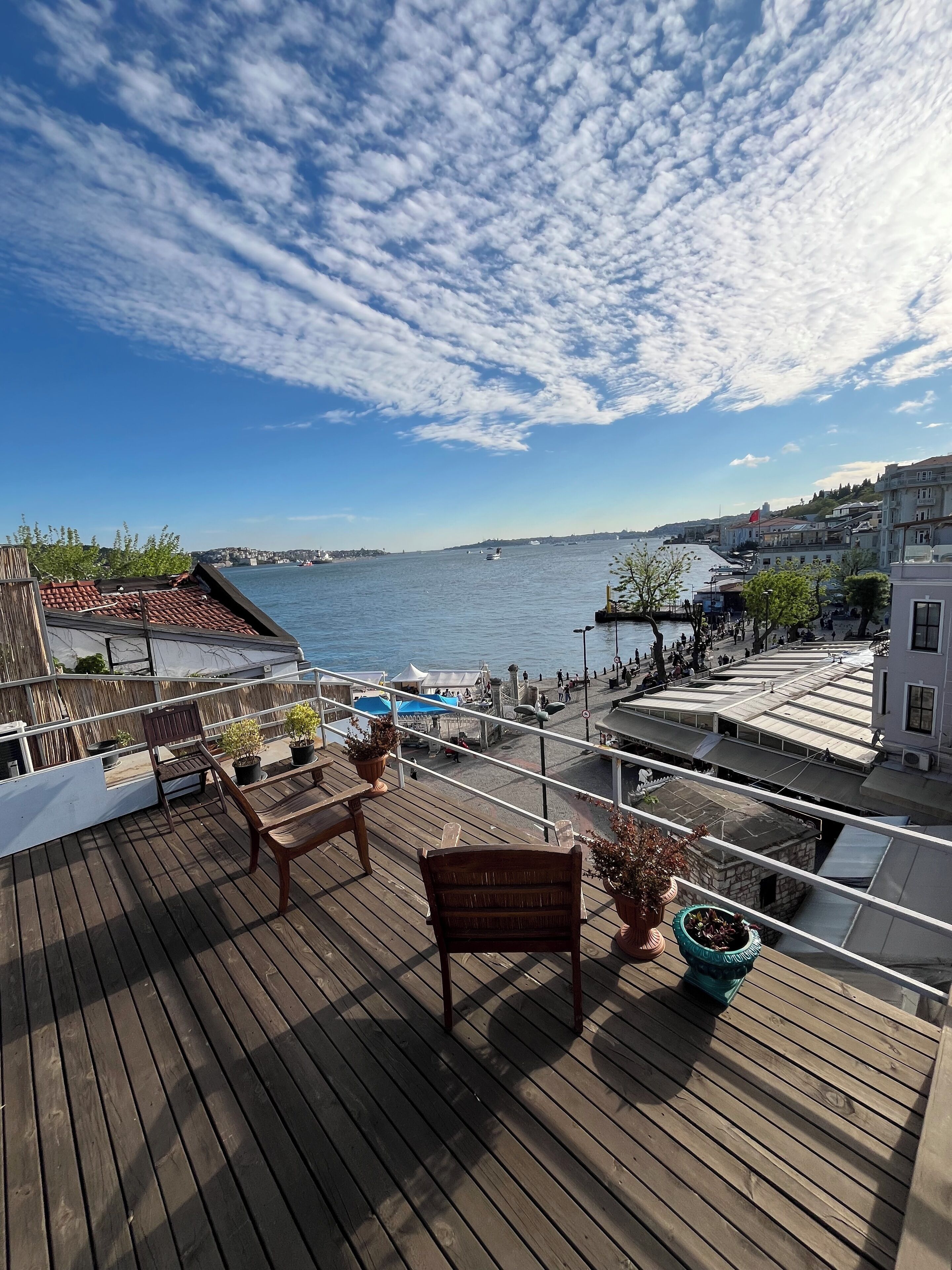 Um deck de madeira com área de estar externa e plantas em vasos, com vista para uma orla com edifícios e céu claro.