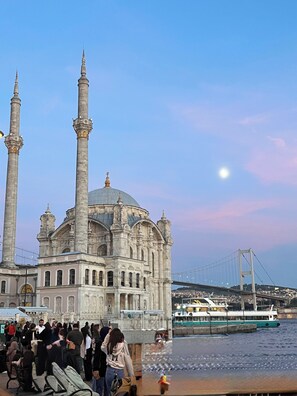 A historic building with multiple minarets and a dome, located near a body of water with a bridge in the background.