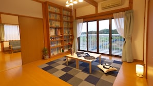 Ein geräumiges Wohnzimmer mit einer großen Bücherwand, einem Schachbrettfußboden, einem Esszimmertisch mit Kissen und Blick durch ein großes Fenster.
