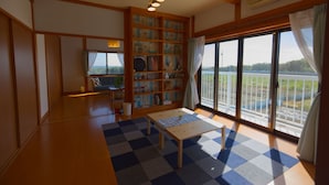 Ein geräumiges Zimmer mit großem Fenster, Holzboden, kariertem Teppich, kleinem Tisch und Bücherregal.