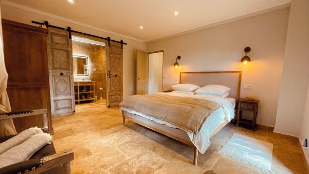 A spacious bedroom with a large bed, a wooden wardrobe, a vanity with a mirror, and a sliding door leading to an adjacent room. - La Réserve Provence (Vaugines)