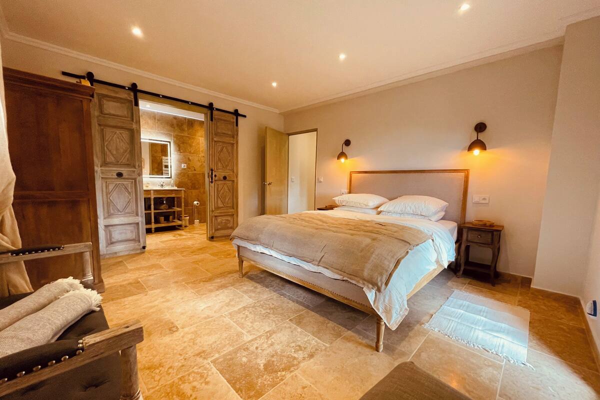 Un dormitorio amplio con una cama grande, un armario de madera, un tocador con espejo y una puerta corrediza que da a una habitación contigua.