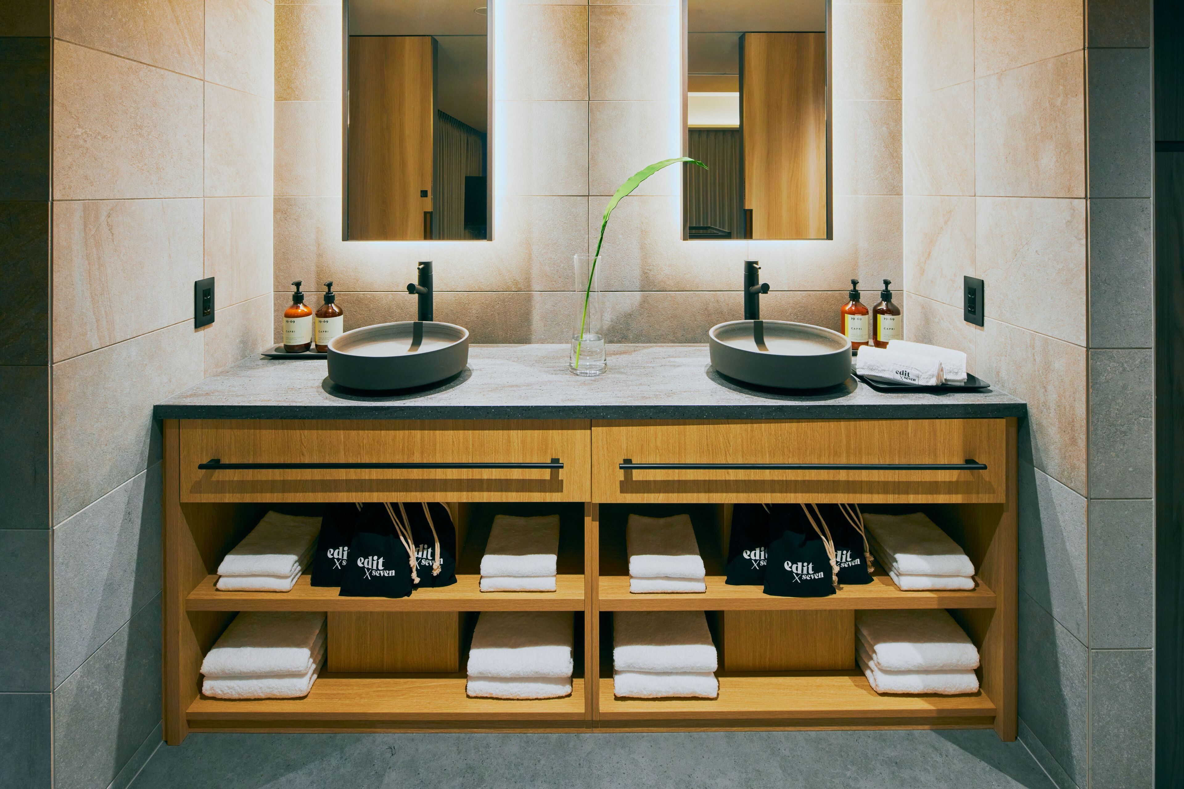 Une salle de bain moderne avec deux lavabos, un miroir et une étagère où sont posées des serviettes et une plante.