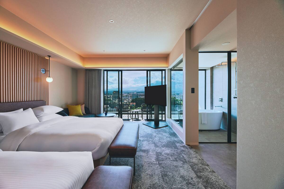 Ein Hotelzimmer mit einem großen Bett, einer Bank, einem Fernseher und einem Balkon mit Blick auf die Stadt.
