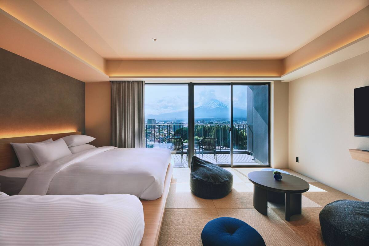 Ein Hotelzimmer mit zwei Betten, einem kleinen Tisch, einem Sitzsack und einem großen Fenster mit Blick auf Stadt und Berge.
